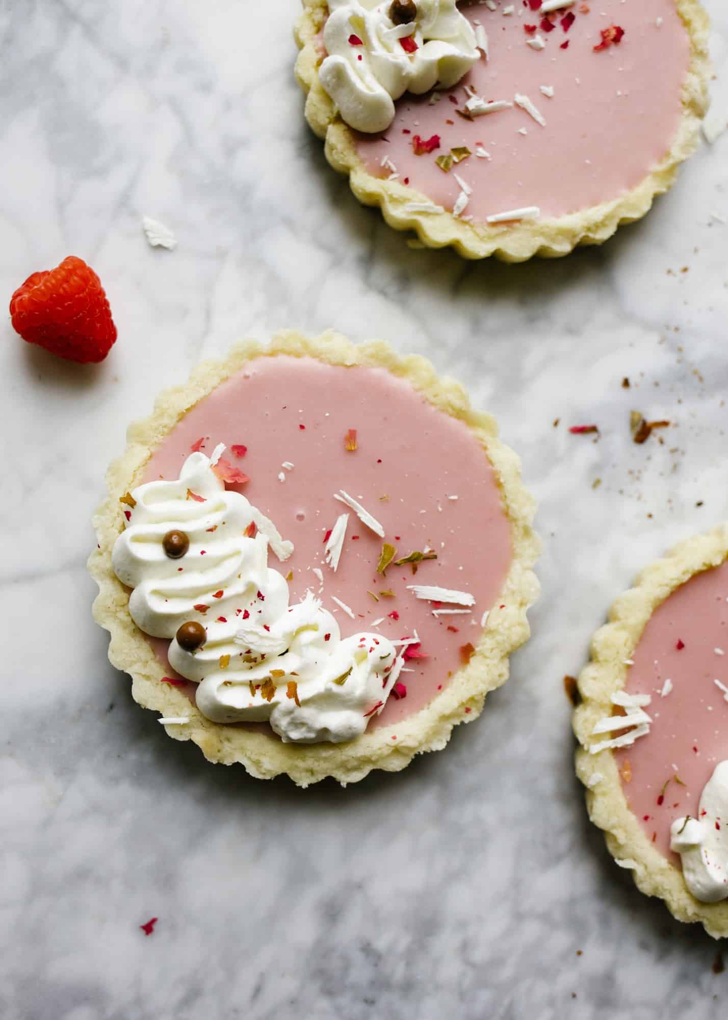 White Chocolate Raspberry Tarts by Wood and Spoon. These are white chocolate and raspberry ganache tarts topped with whipped cream and filled into a shortbread press in crust. These pink tarts are cute for Valentine's Day or any other pinkies out kind of event. Find the recipe and how to for these simple make ahead treats on thewoodandspoon.com