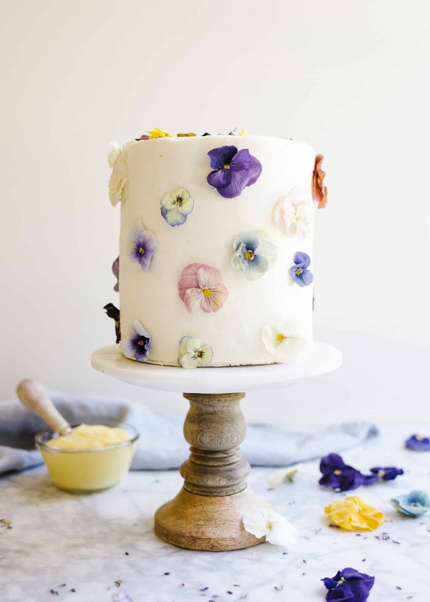 Lavender Cake with Lemon Curd and Creamy Vanilla Frosting by Wood and Spoon blog. This is a floral cake decorated with edible dried flowers and filled with a homemade lemon curd. This sturdy lavender cake is coated in fluffy American whipped buttercream. Learn how to make this beautiful layer cake for the spring on thewoodandspoon.com