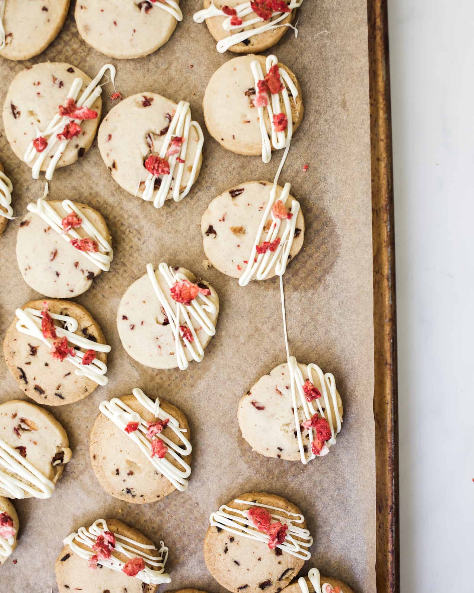 Strawberry Shortbread Cookies by Wood and Spoon blog. These are simple buttery cookies with bits of dried strawberries throughout. A while chocolate drizzle finishes off the crisp cookies that make a terrific treat to go with coffee or tea! Find out how to make freeze dried fruit cookies on thewoodandspoon.com